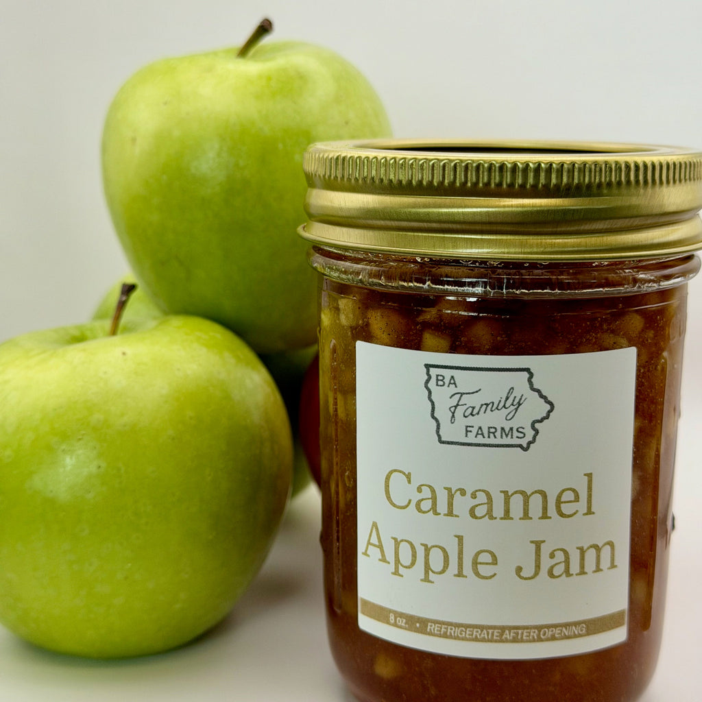 A jar of BA Family Farms Caramel Apple Jam with a gold lid and white label, shown next to two green apples on a white background.