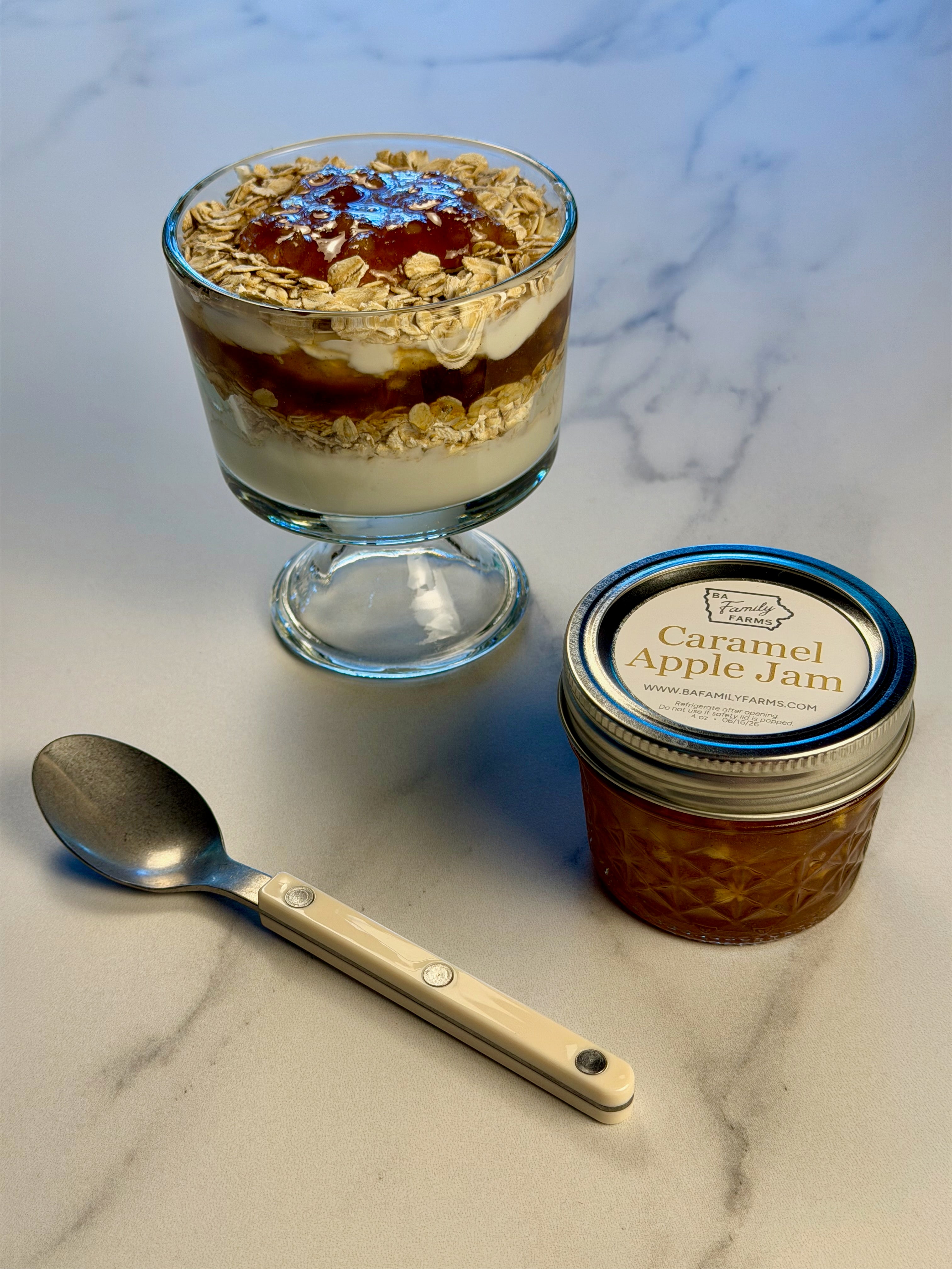 A small jar of Caramel Apple Jam sits on a light marble surface next to a spoon and a dessert glass filled with layers of yogurt, fruit, and oats. The jar label reads 'Caramel Apple Jam'.