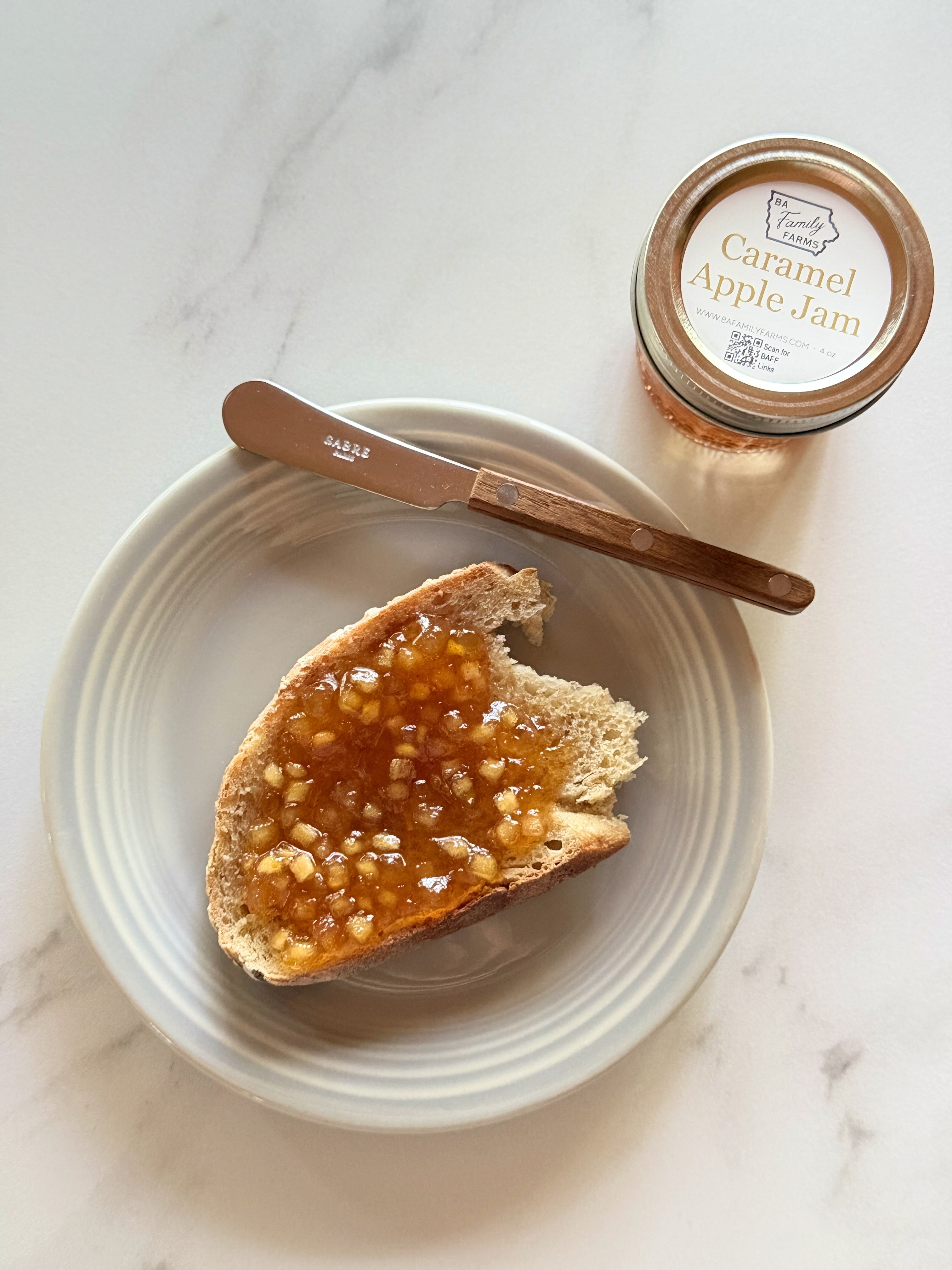 A jar of Caramel Apple Jam sits on a white marble surface next to a plate with a slice of toast topped with the jam and a wooden butter knife.