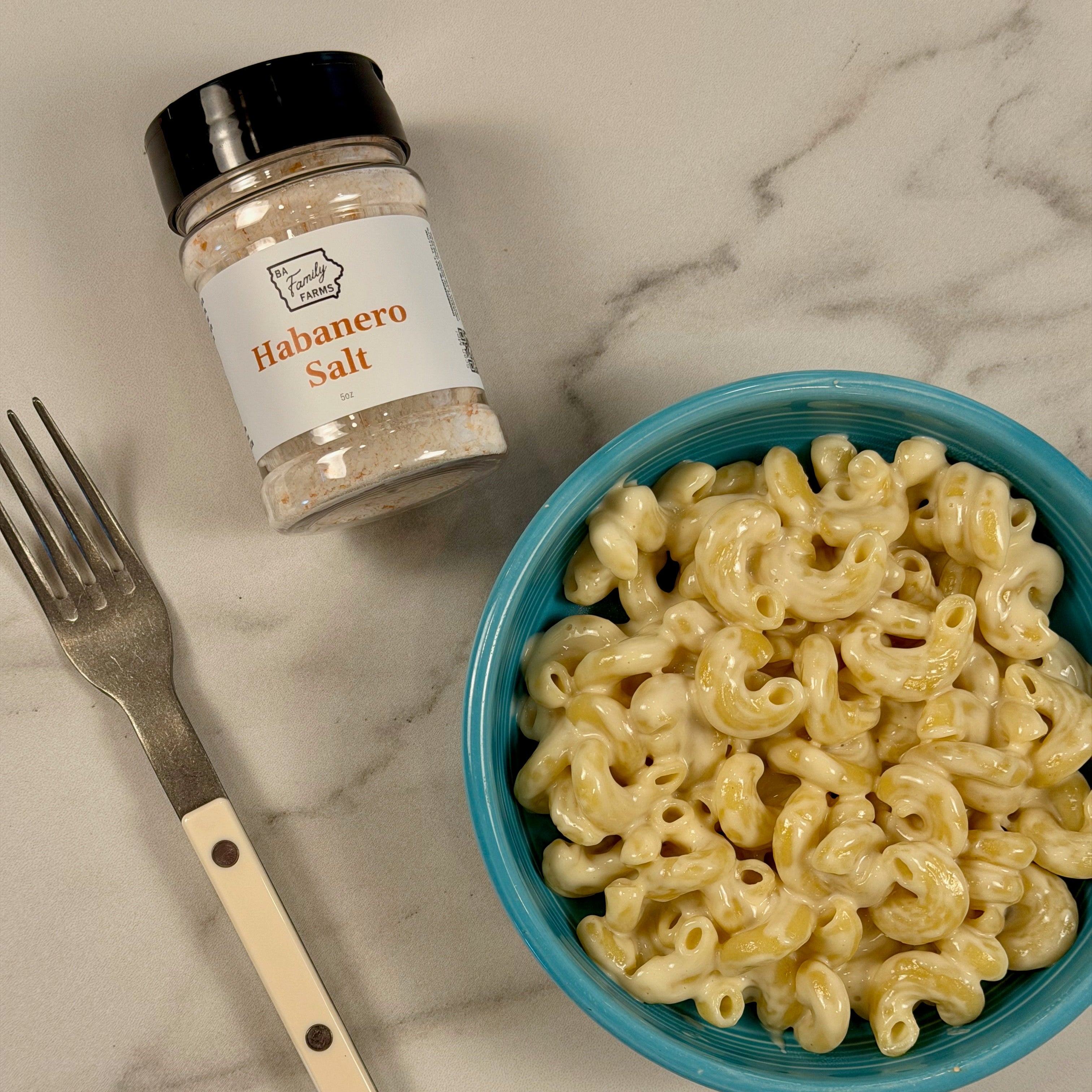 A bowl of macaroni pasta with a blue rim, a small jar labeled Habanero Salt, and a metal fork with a cream handle, arranged on a light marble surface.