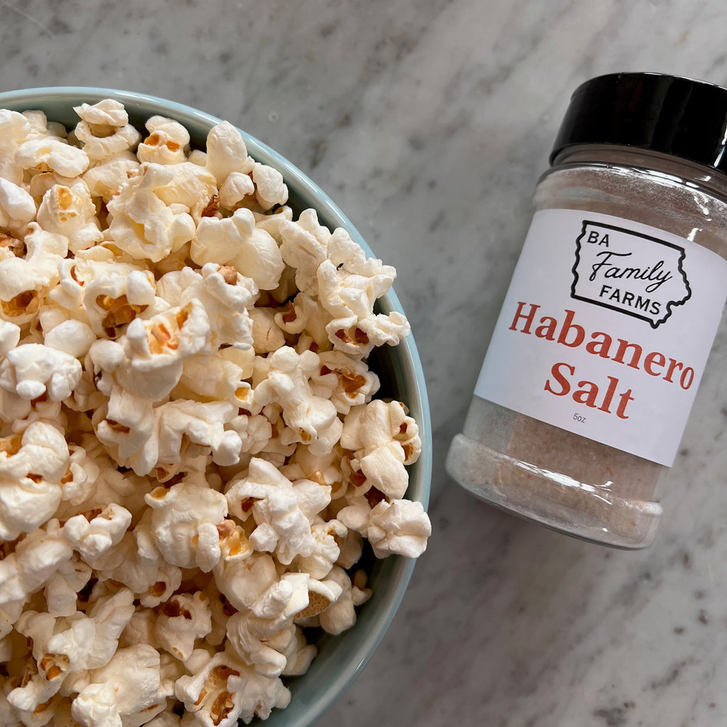 A bowl of popcorn next to a spice jar labeled 'BA Family Farms Habanero Salt' on a light gray marble surface.