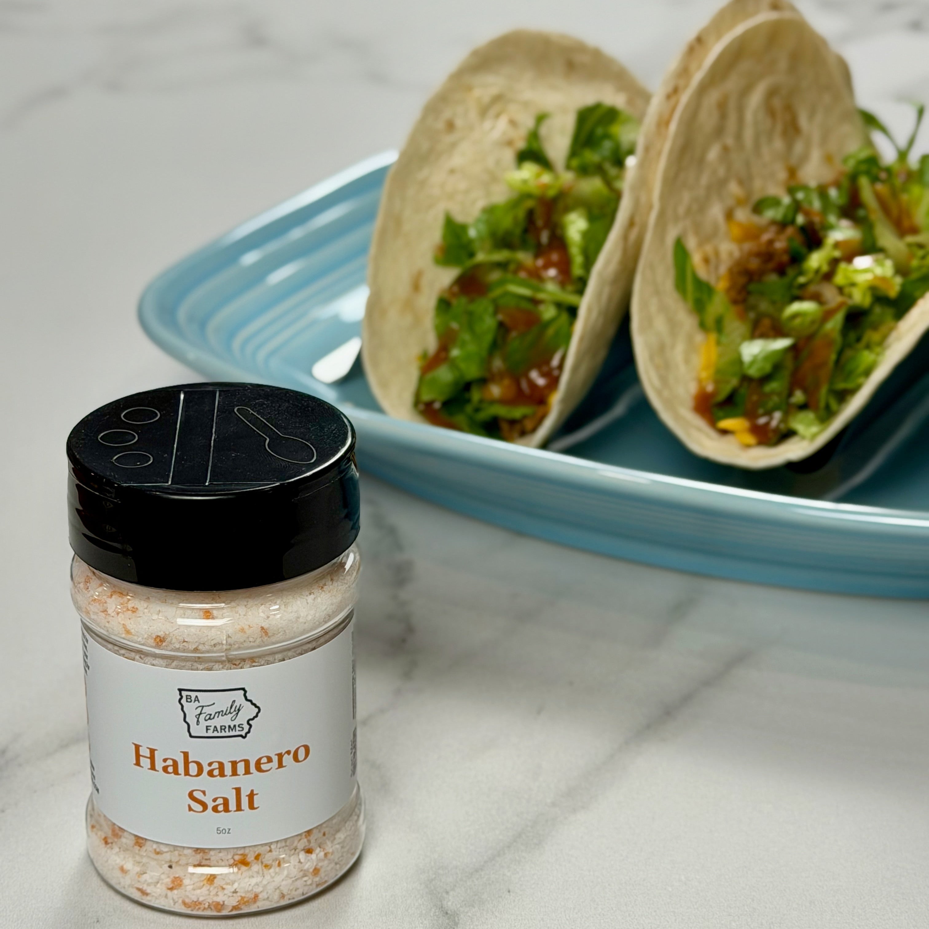 A shaker bottle of BA Family Farms Habanero Salt in the foreground on a white marble surface, with two tacos on a light blue plate blurred in the background.