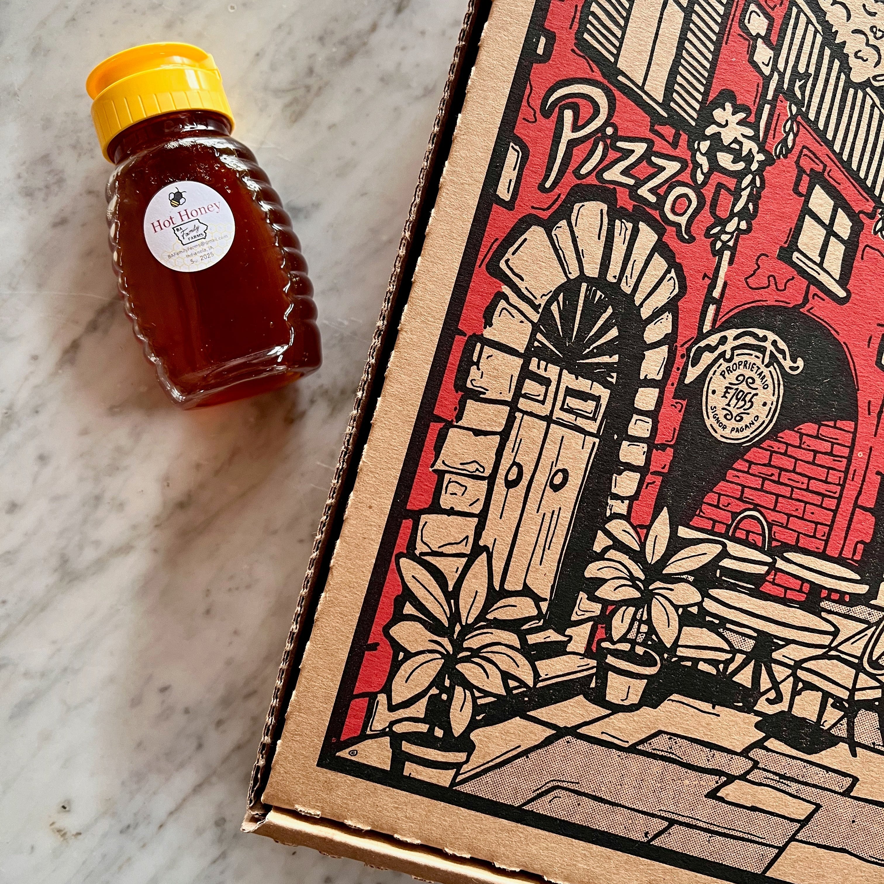 A BA Family Farms Hot Honey bottle with a yellow cap sits next to a cardboard pizza box featuring a red, black, and beige illustration of a pizza oven and the word 'Pizza' on a light marble surface.