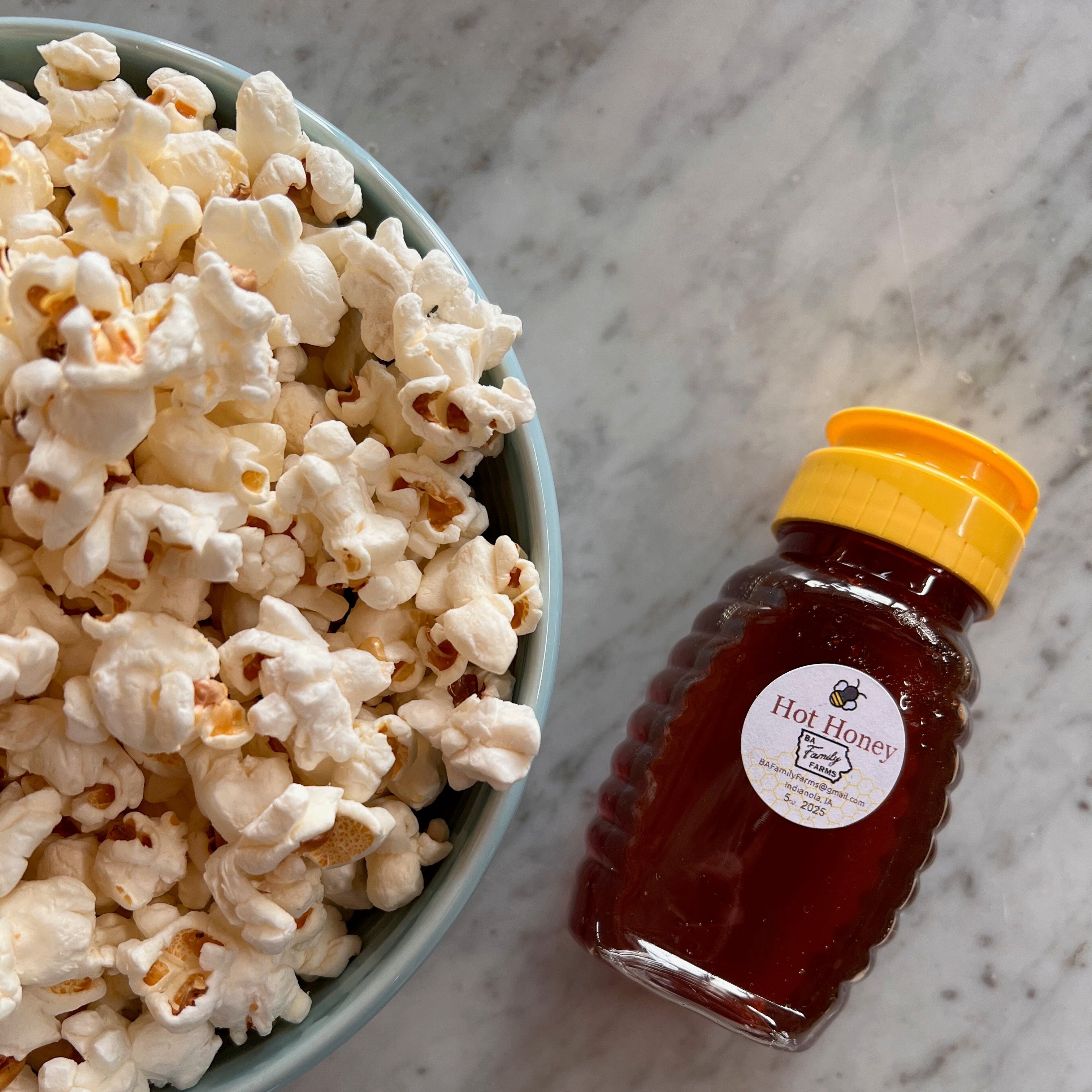 A bottle of BA Family Farms hot honey with a yellow cap and a round white label sits on a light marble surface beside a bowl of popcorn.