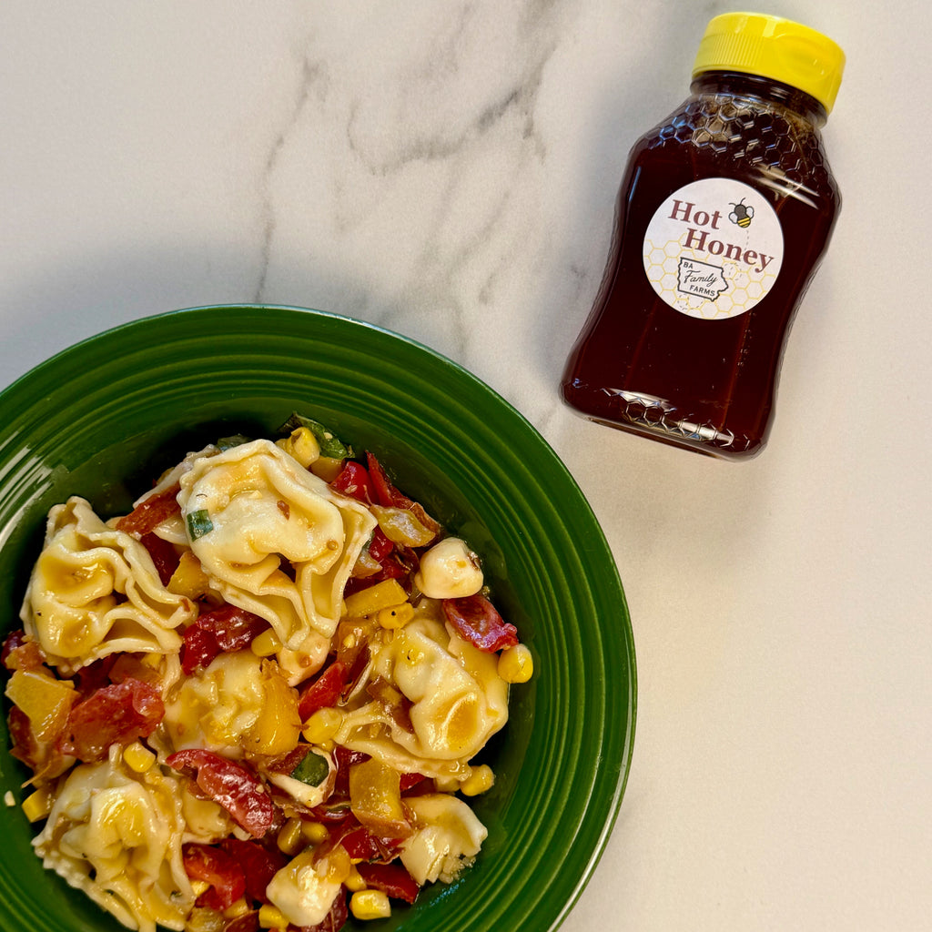 A green bowl filled with a pasta salad containing tortellini, corn, and pieces of cured meat, placed on a white marble surface next to a bottle of BA Family Farms hot honey.