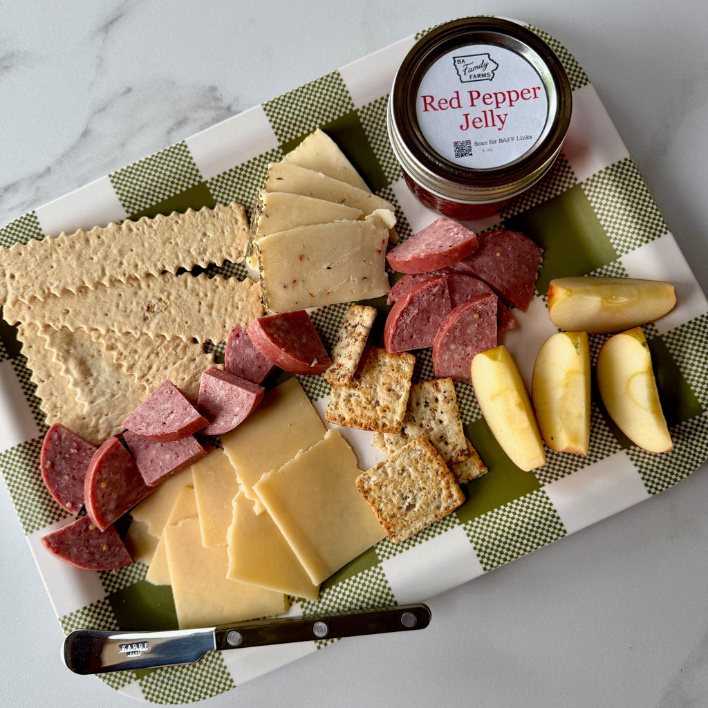 A charcuterie board styled with crackers, sliced cheeses, salami, apple wedges, and a small jar of BA Family Farms Red Pepper Jelly on a green-and-white checkered cloth over a marble surface.