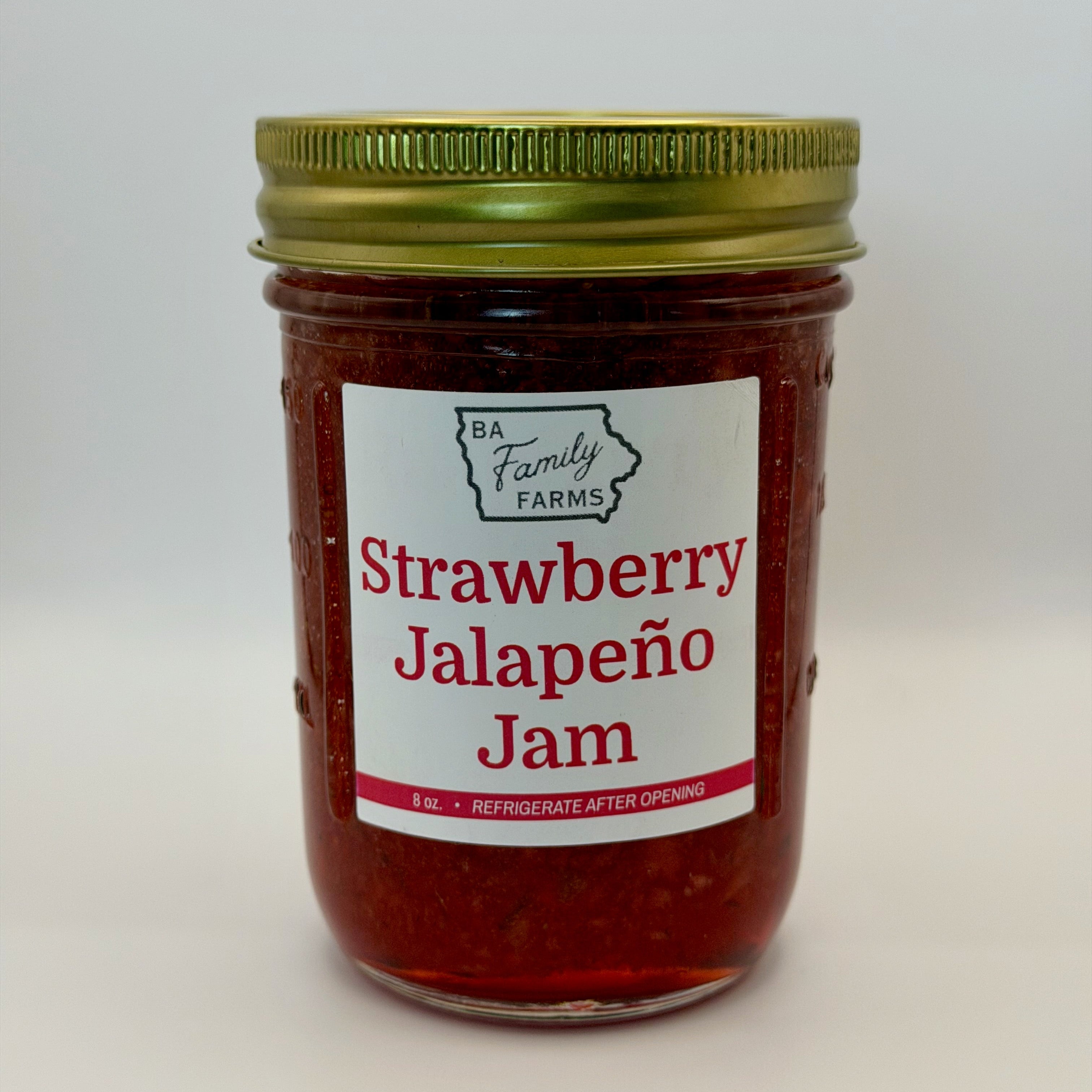 A glass jar of Strawberry Jalapeño Jam with a white label and a gold metal lid, photographed against a plain light gray background.