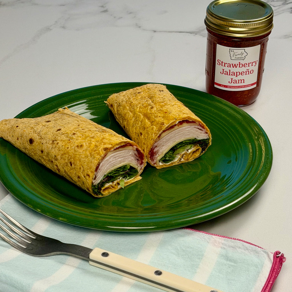 A green plate holding two cut wrap sandwiches with visible filling, a jar of Strawberry Jalapeño Jam behind it, and a fork on a light blue striped napkin on a white marble surface.