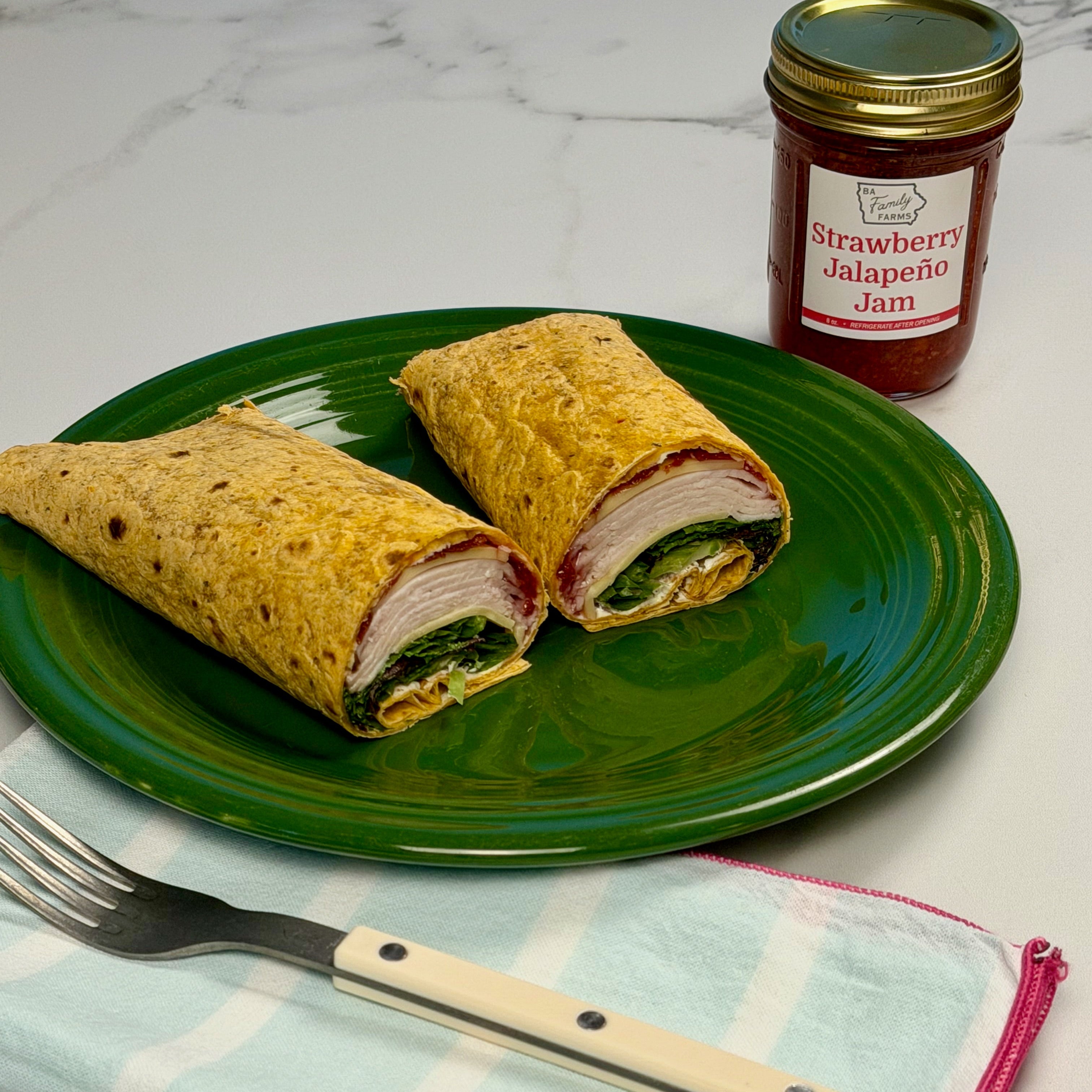 A green plate holding two cut wrap sandwiches with visible filling, a jar of Strawberry Jalapeño Jam behind it, and a fork on a light blue striped napkin on a white marble surface.