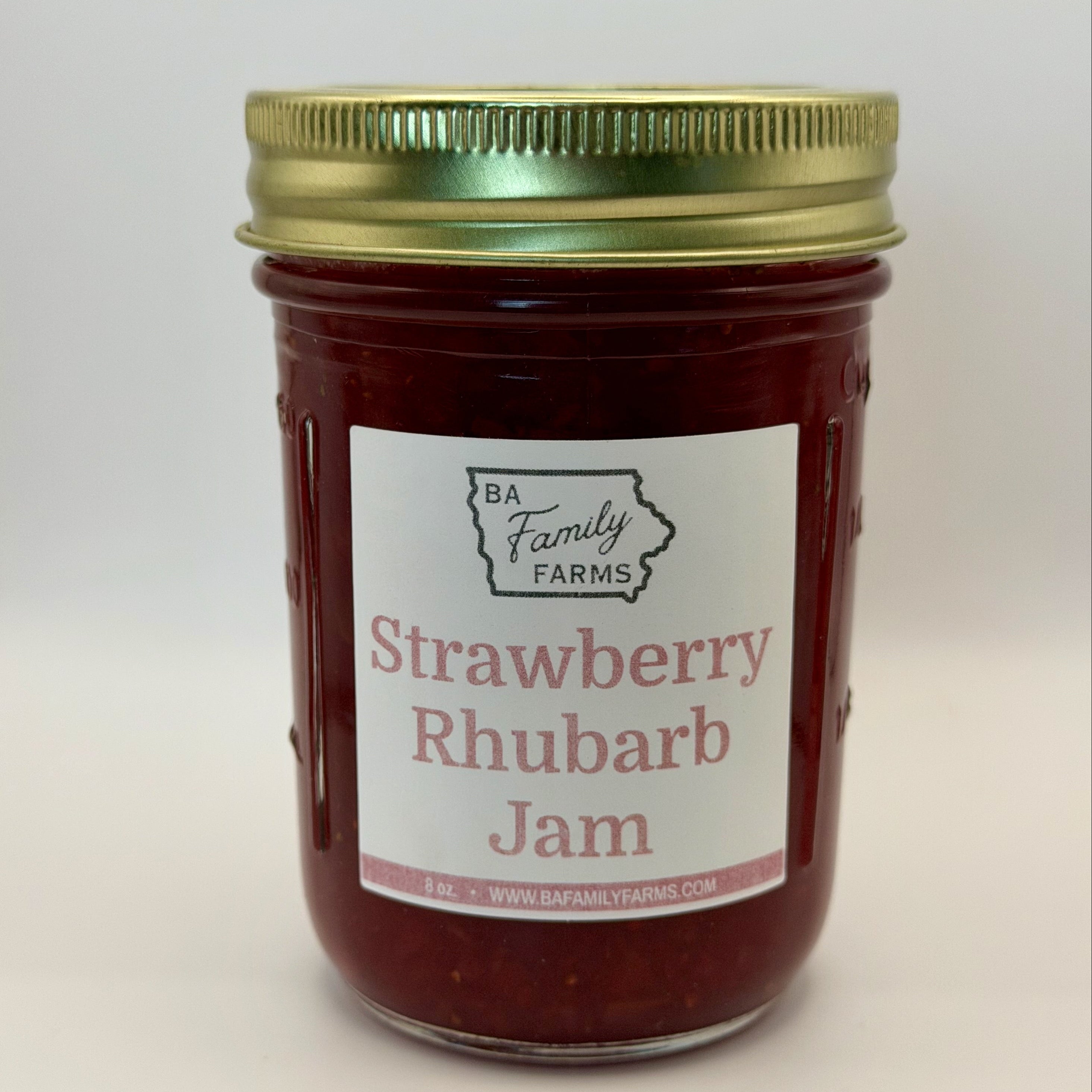 A glass jar of BA Family Farms Strawberry Rhubarb Jam with a white label and a gold metal lid, set against a plain light gray background.