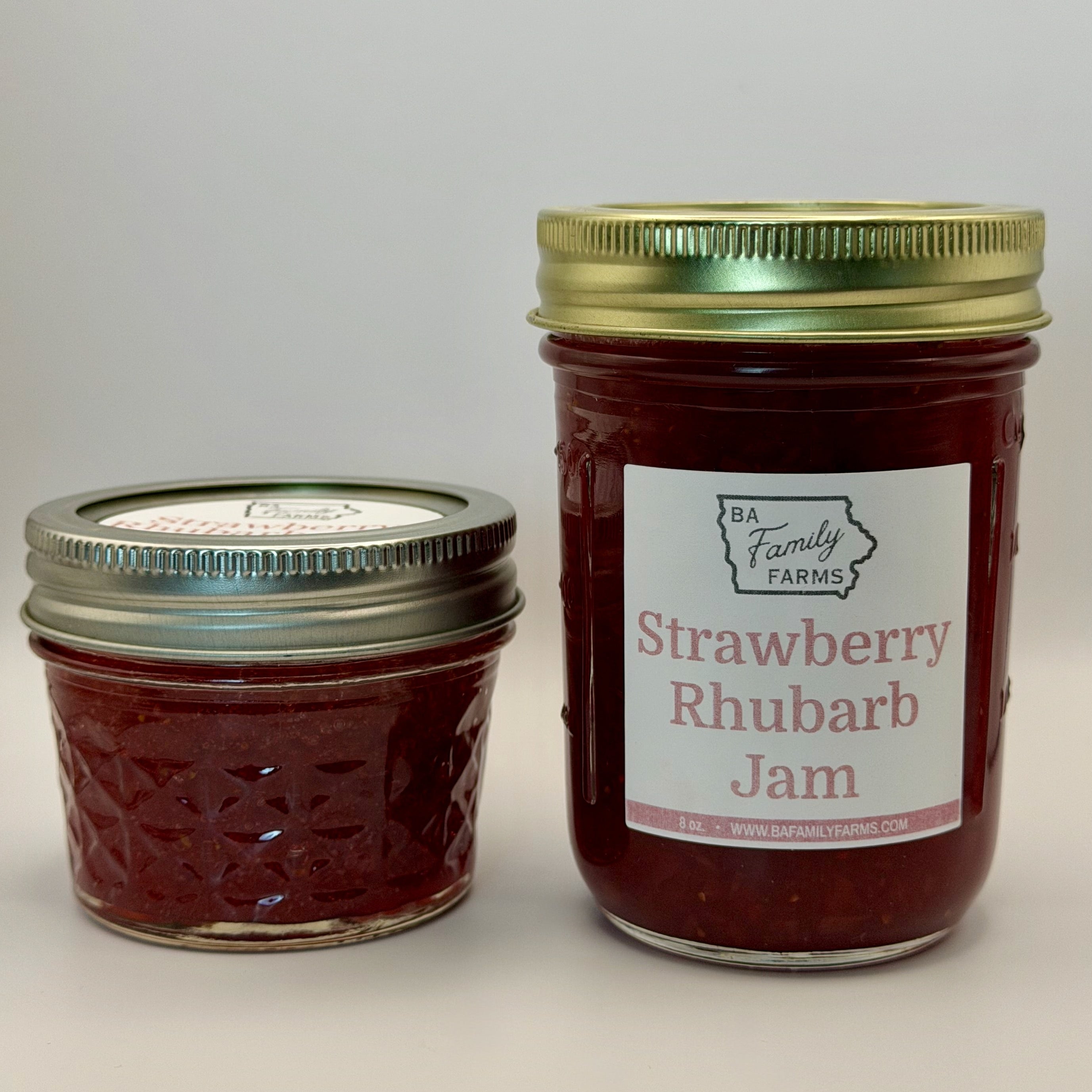 Two glass jars of BA Family Farms Strawberry Rhubarb Jam on a light gray background, with one small jar and one larger jar. The larger jar has a white label with red text and a gold metal lid, while the smaller jar has a silver metal lid.