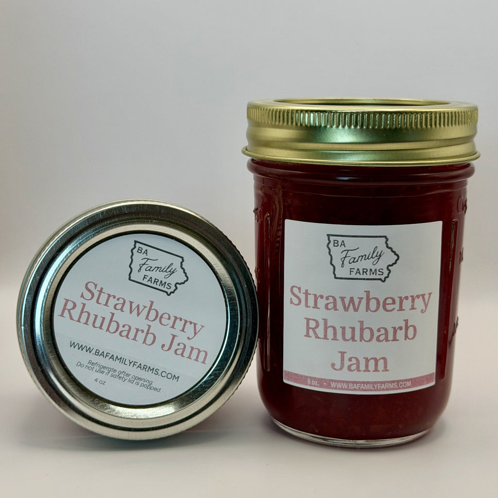 A jar of Strawberry Rhubarb Jam by BA Family Farms stands upright next to its lid on a light gray background. The jar contains deep red jam and has a gold metal lid, while the lid in front shows the product label.