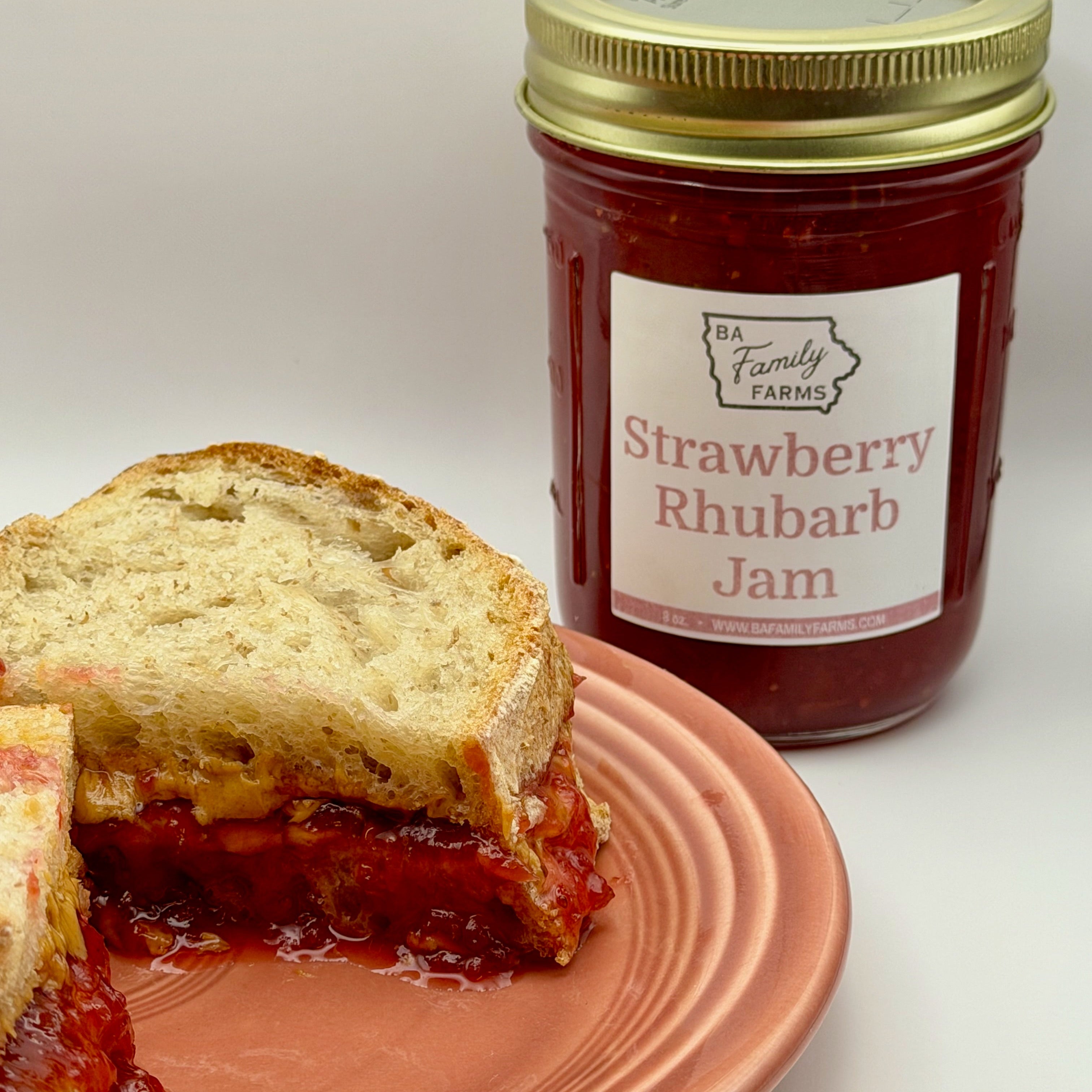 A jar of BA Family Farms Strawberry Rhubarb Jam stands behind a cut sandwich filled with jam on a peach-colored plate against a light gray background.